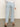 Person stands indoors facing away, wearing Sanctuary Clothing's district column ankle jean in blue mirage with a white long-sleeve top and brown sandals. The background is plain and neutral.