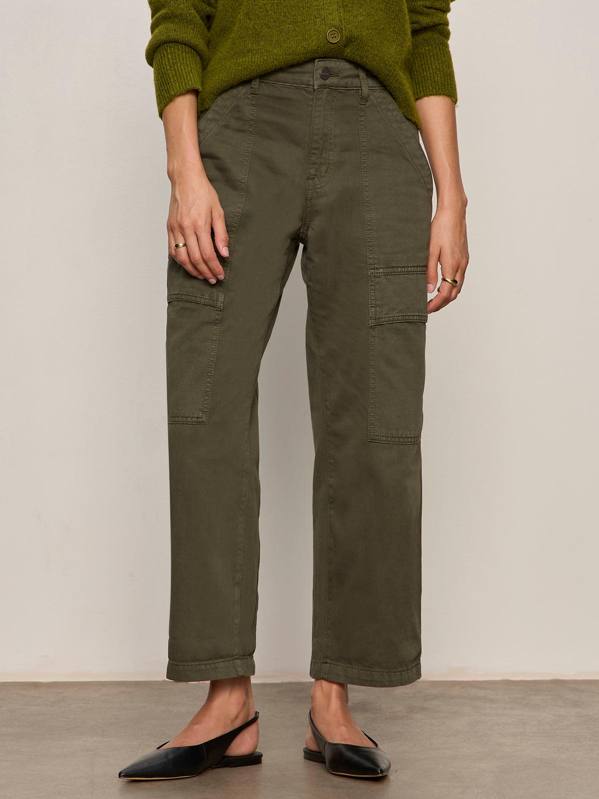 A person wearing Sanctuary Clothing's boyfriend utility pant kalamata and a green sweater with rolled-up sleeves, styled with black pointed-toe slingback shoes, stands against a plain light background.