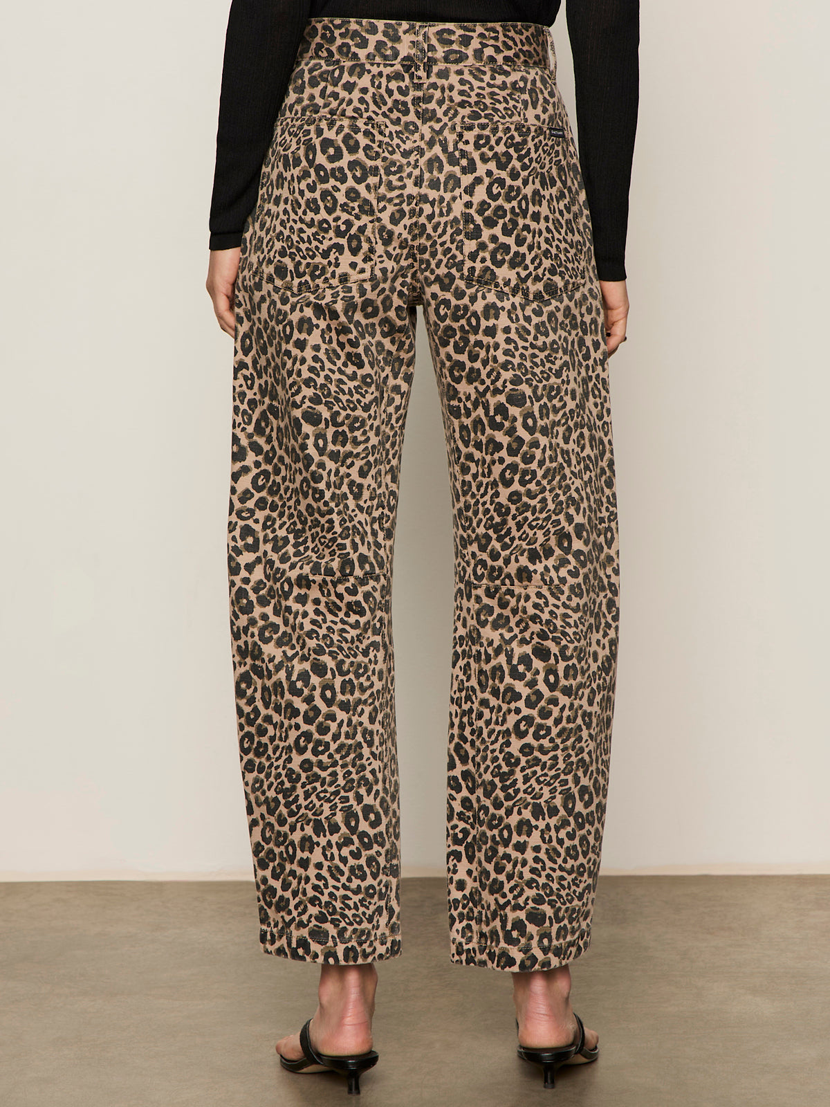 A person stands on a concrete floor wearing Sanctuary Clothing's boyfriend utility high rise pant in French Kitty, paired with a black long-sleeve top and black heeled sandals. The photo captures the back view of the outfit.