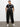 A woman stands against a white wall, confidently posing with her hands in the pockets of black sahara tapered pants from Sanctuary Clothing's inclusive collection, styled with a black and white printed T-shirt and white sneakers.