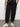 A person wearing Sanctuary Clothing™s sahara tapered pant black inclusive collection with pockets and white sneakers stands on a gray floor by a plain white wall; only their lower half is shown, hands in pockets.