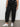 A person wearing Sanctuary Clothing's sahara tapered pant black from the inclusive collection stands against a plain wall, arms relaxed by their sides and upper body partially out of frame.