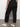 A person stands indoors facing away, wearing Sanctuary Clothing™s sahara tapered pant black inclusive collection with back pockets, a black-and-white patterned top, and white Veja sneakers against a plain neutral background.