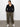 A woman stands against a plain white wall, wearing the Sanctuary Clothing sahara tapered pant in burnt olive from the inclusive collection, paired with a black long-sleeve button-up top and white sneakers. Her hands are in her pockets as she looks slightly to the side.
