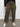 Only the lower torso and legs are visible as someone stands against a plain white wall, wearing Sanctuary Clothing's sahara tapered pant in burnt olive from the inclusive collection, paired with a long-sleeve black top and white sneakers.