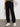Only the lower half of a person is visible as they stand against a white wall, wearing Sanctuary Clothing's sahara tapered pant in black.