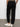 A person stands on a gray floor facing away from the camera, wearing Sanctuary Clothing™s sahara tapered pant in black, a khaki shirt, and white Adidas sneakers with three stripes; hands are in their pockets.