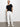 A person stands against a plain white wall, wearing a black short-sleeve t-shirt, white sahara tapered pant chalk by Sanctuary Clothing, and white sneakers, with one hand in a pocket and a neutral expression.