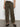 A person stands against a plain wall, wearing Sanctuary Clothing's sahara tapered pant in burnt olive and a white t-shirt. Their black woven shoes are visible below the pants, with arms resting by their sides and only their torso down shown.