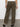 A person faces away, wearing Sanctuary Clothing's sahara tapered pant in burnt olive with back pockets and a white shirt. They have black woven shoes and stand on a concrete floor against a plain white wall.