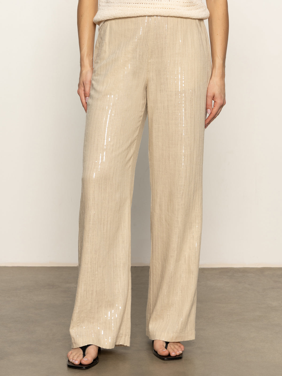 Someone wearing the Sanctuary Clothing coastal pant sand sequin stripe—light beige, relaxed-fit pants with subtle vertical sequin stripes—pairs them with black open-toe sandals, standing on a smooth floor against a plain light background.