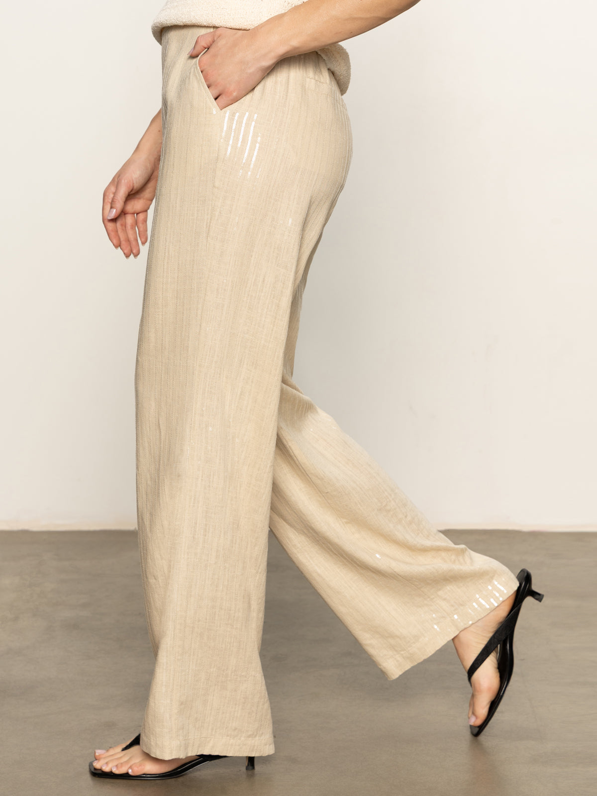 A person wearing Sanctuary Clothing’s coastal pant sand sequin stripe and black heeled sandals walks indoors on a polished floor, one hand in their pocket. The image is cropped above the waist.