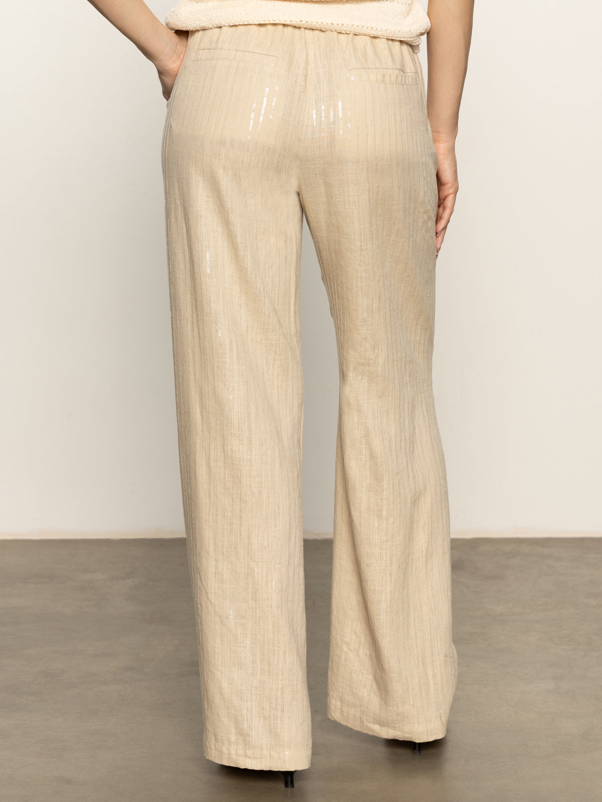 A person stands indoors on a gray floor, back to the camera, wearing Sanctuary Clothing's coastal pant sand sequin stripe—light beige, shiny, wide-leg pants—paired with black heeled shoes.