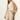 A woman with long blonde hair wears a sleeveless cream knit top and Sanctuary Clothing’s coastal pant sand sequin stripe—high-waisted, light beige pants with subtle shine—standing against an off-white wall, one hand in her pocket.
