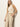 A woman with long blonde hair wears a sleeveless cream knit top and Sanctuary Clothing’s coastal pant sand sequin stripe—high-waisted, light beige pants with subtle shine—standing against an off-white wall, one hand in her pocket.