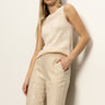 A woman with long blonde hair wears a sleeveless cream knit top and Sanctuary Clothing’s coastal pant sand sequin stripe—high-waisted, light beige pants with subtle shine—standing against an off-white wall, one hand in her pocket.