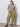 A woman with long dark hair poses with one hand in her pocket, wearing Sanctuary Clothing's area standard rise pant bamboo extended sizing in olive green, paired with a black-and-white striped tee and black sandals against a plain wall.