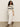 A woman with curly hair walks on a beige floor, wearing a cream knit sweater with black stripes, Sanctuary Clothing's area standard rise pant in bleached white, and black sandals. The background is plain and light-colored.
