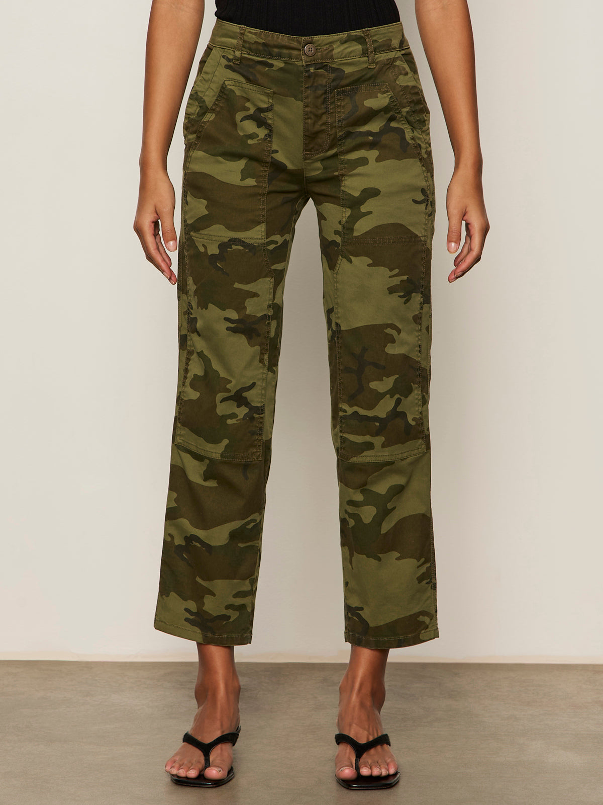 A person models the Sanctuary Clothing rio pant rambler camo cargo pants with black heeled sandals, standing against a beige background with arms relaxed at their sides and upper body not shown.