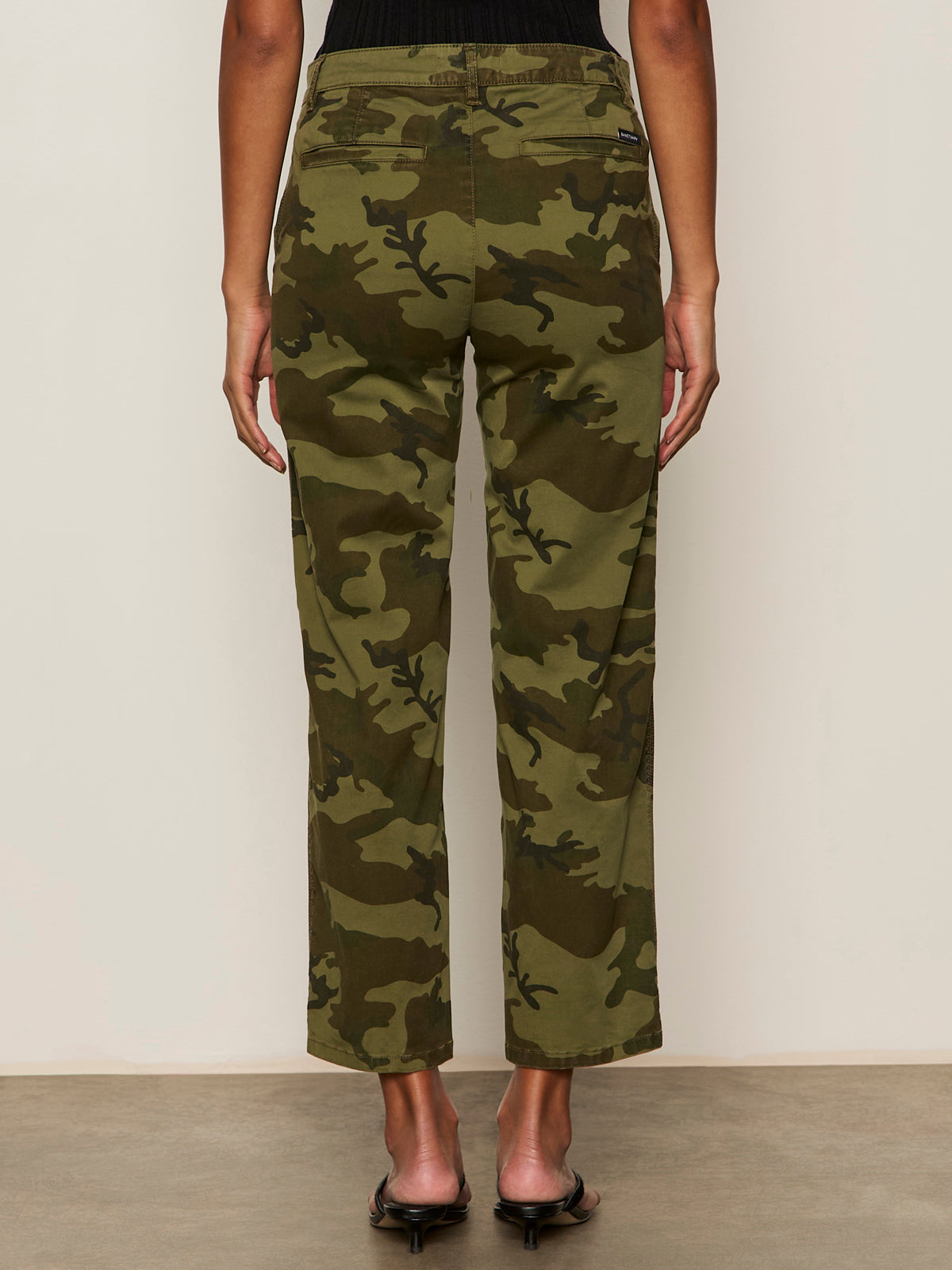 A model faces away, showing the back of Sanctuary Clothing's rio pant rambler camo against a plain light wall, paired with black heeled sandals.