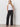 A woman with long, wavy blonde hair and a relaxed expression stands against a white wall wearing Sanctuary Clothing's Cruiser Full Length Black pants, a white sleeveless halter top, and black heeled sandals.