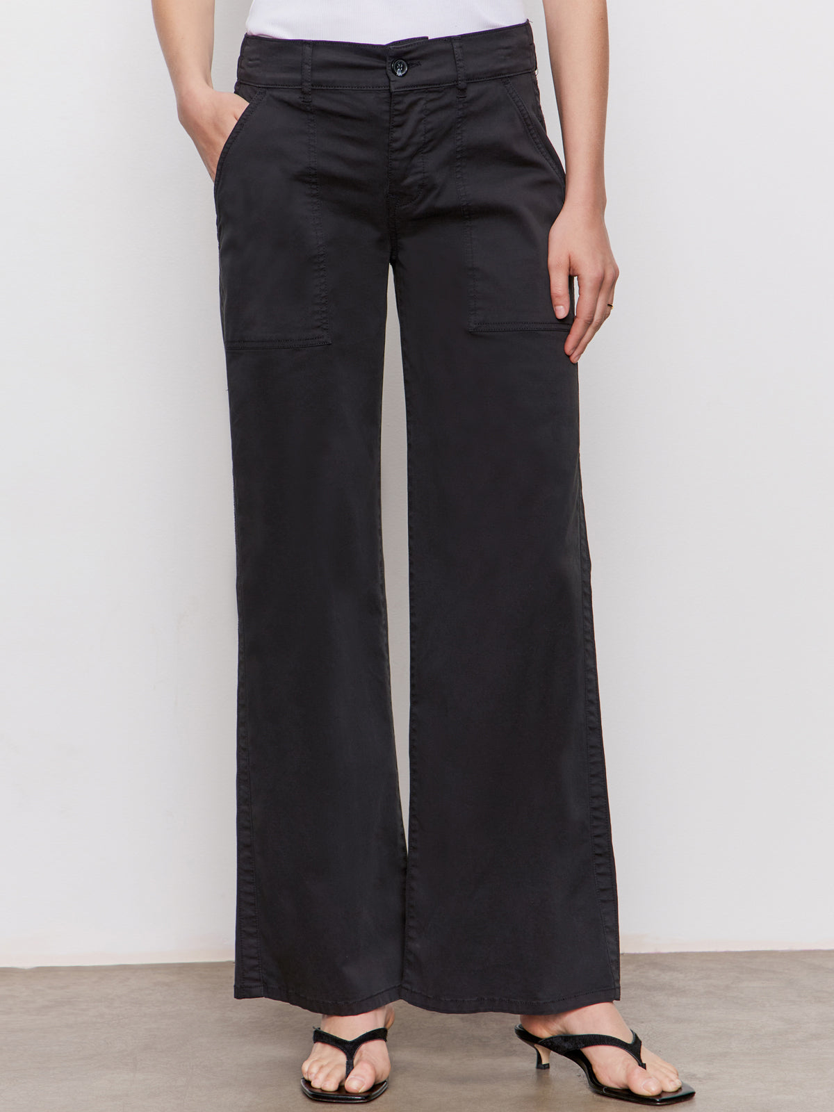 A person stands against a white wall wearing Sanctuary Clothing's cruiser full length black trousers with front patch pockets and black heeled sandals, hands in pockets, lower body only visible.