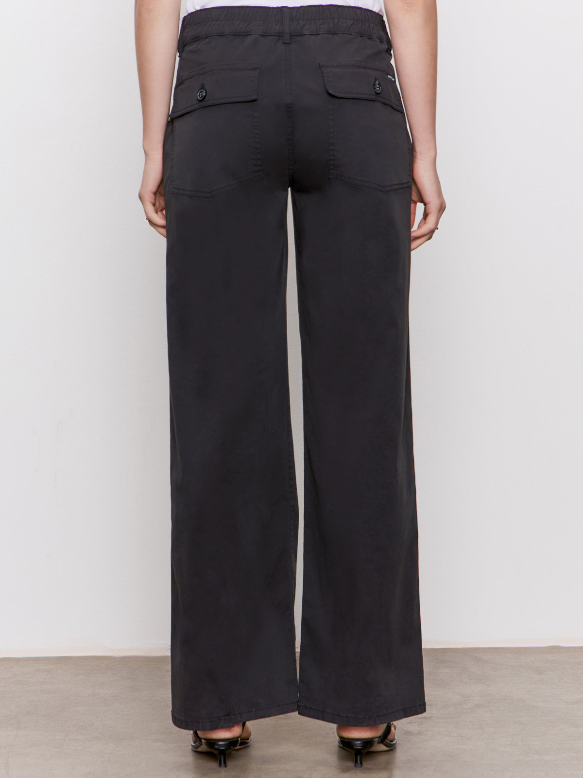 Back view of a person wearing Sanctuary Clothing's cruiser full length black pants, featuring high waist, wide legs, two large back pockets with button flaps, and an elastic waistband, shown on a neutral-colored floor.