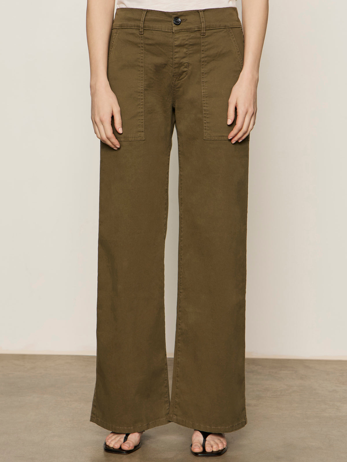 A person wearing the Sanctuary Clothing cruiser full length mossy green pants is shown from the waist down, standing against a plain beige background. The pants have front patch pockets and a wide-leg design.