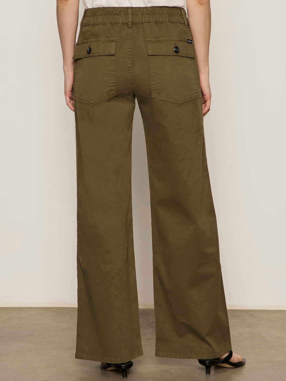 Back view of a person wearing Sanctuary Clothing's cruiser full length mossy green wide-leg pants with back pockets and black button closures, standing on a light brown floor against an off-white wall.