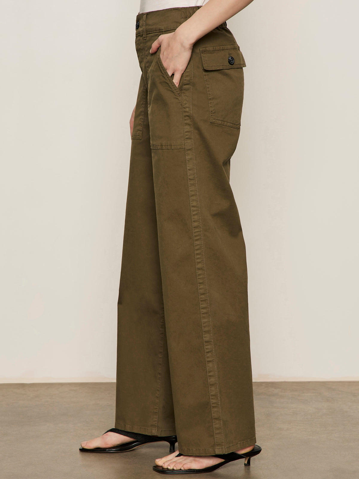 A person wearing Sanctuary Clothing's cruiser full length mossy green pants, featuring a high-waisted, wide-leg design with side pockets and black heeled sandals, stands against a neutral background.