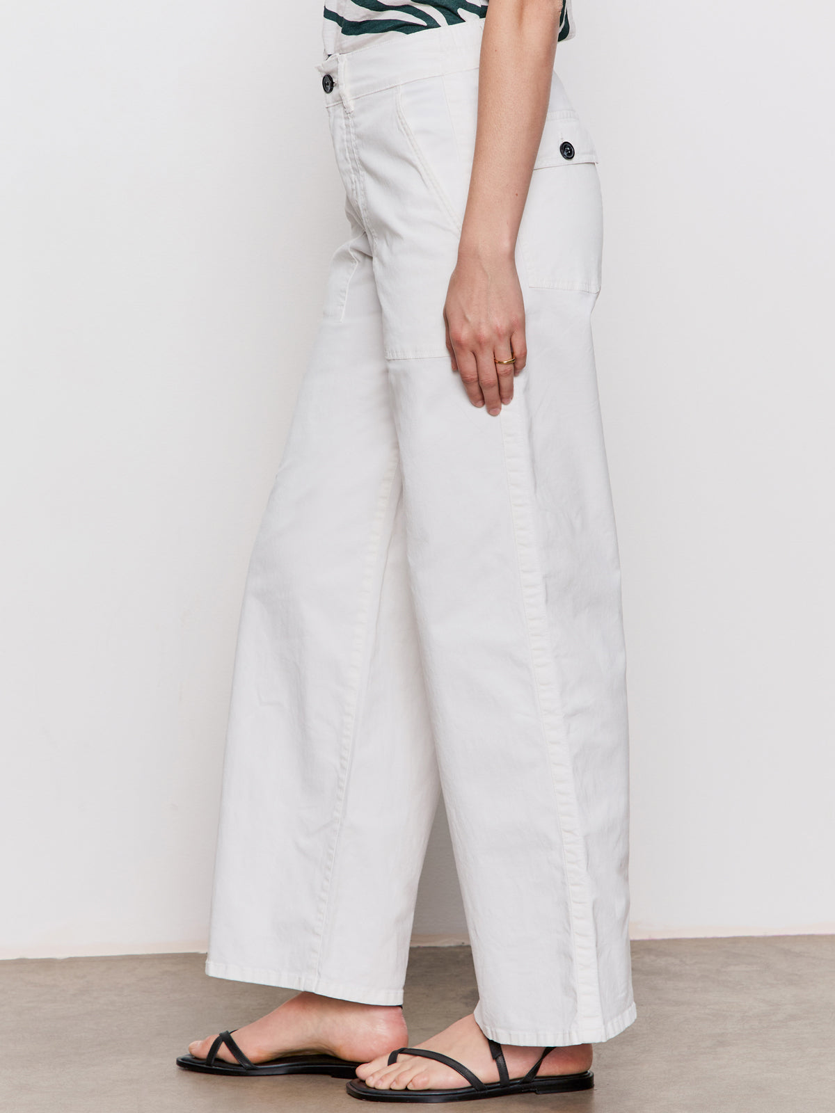 A person models the Sanctuary Clothing cruiser full length powdered sugar high-waisted, wide-leg pants and black sandals, standing against a plain background with their lower body visible and arm relaxed at their side.