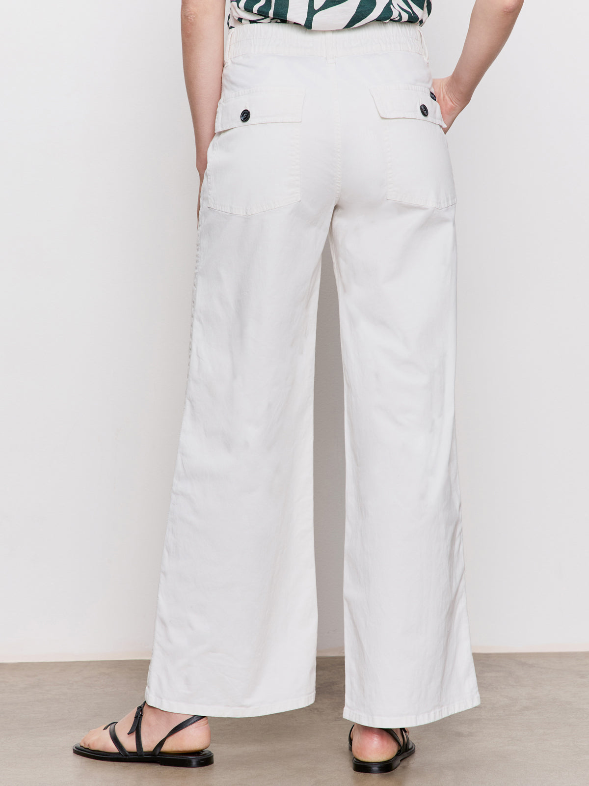 A person stands indoors facing away from the camera, wearing Sanctuary Clothing's Cruiser Full Length Powdered Sugar wide-leg pants with back patch pockets and black buttons, black flat sandals, and a partially visible green and white patterned top.
