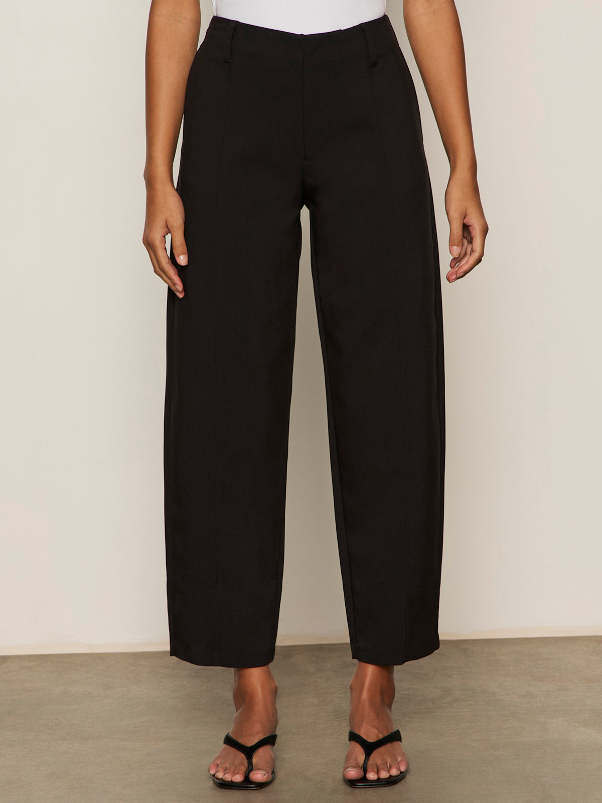 A person wearing Sanctuary Clothing's polished barrel pant in black with black heeled sandals, standing against a light wall and concrete floor. Only the lower half of the body is visible.