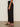 A person stands sideways wearing Sanctuary Clothing's polished barrel pant in black, paired with a white top and black heeled sandals on a smooth, neutral floor against an off-white wall.
