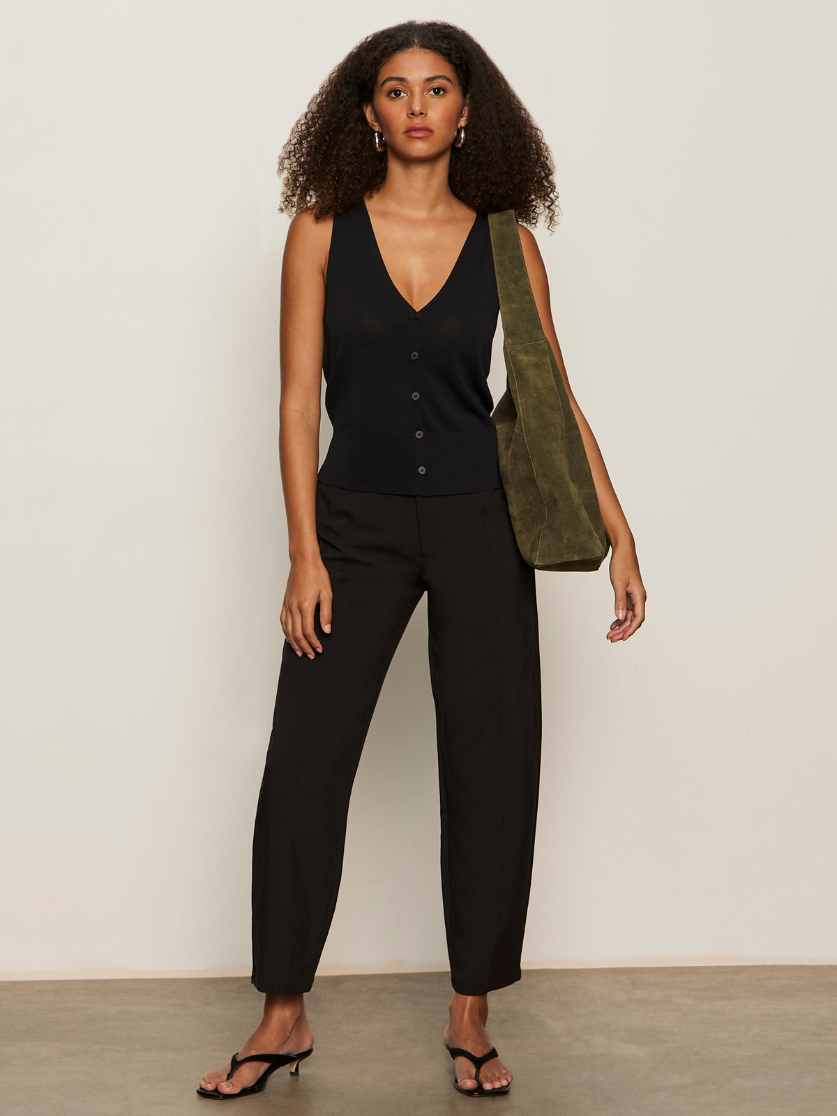 A woman with curly hair wears a sleeveless black V-neck top, Sanctuary Clothing polished barrel pant in black, and black heeled sandals. She stands against a plain background, holding an olive green shoulder bag.