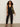 A woman with curly hair wears a sleeveless black V-neck top, Sanctuary Clothing polished barrel pant in black, and black heeled sandals. She stands against a plain background, holding an olive green shoulder bag.