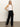 A woman poses confidently against a plain backdrop in Sanctuary Clothing's casual track pant black chalk stripe, styled with a white sleeveless top and black sandals. Her straight blonde hair complements the relaxed look.