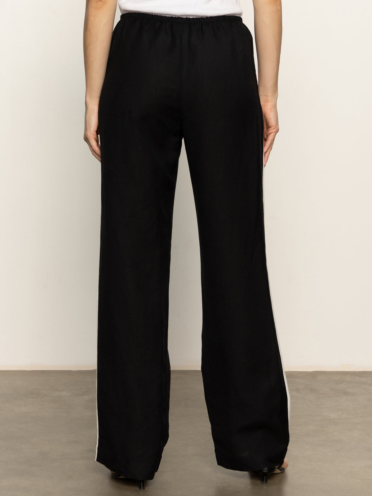 A person wearing Sanctuary Clothing’s casual track pant in black chalk stripe, featuring an elastic waistband and loose fit, is viewed from the back while standing on a smooth floor against a plain light-colored wall.
