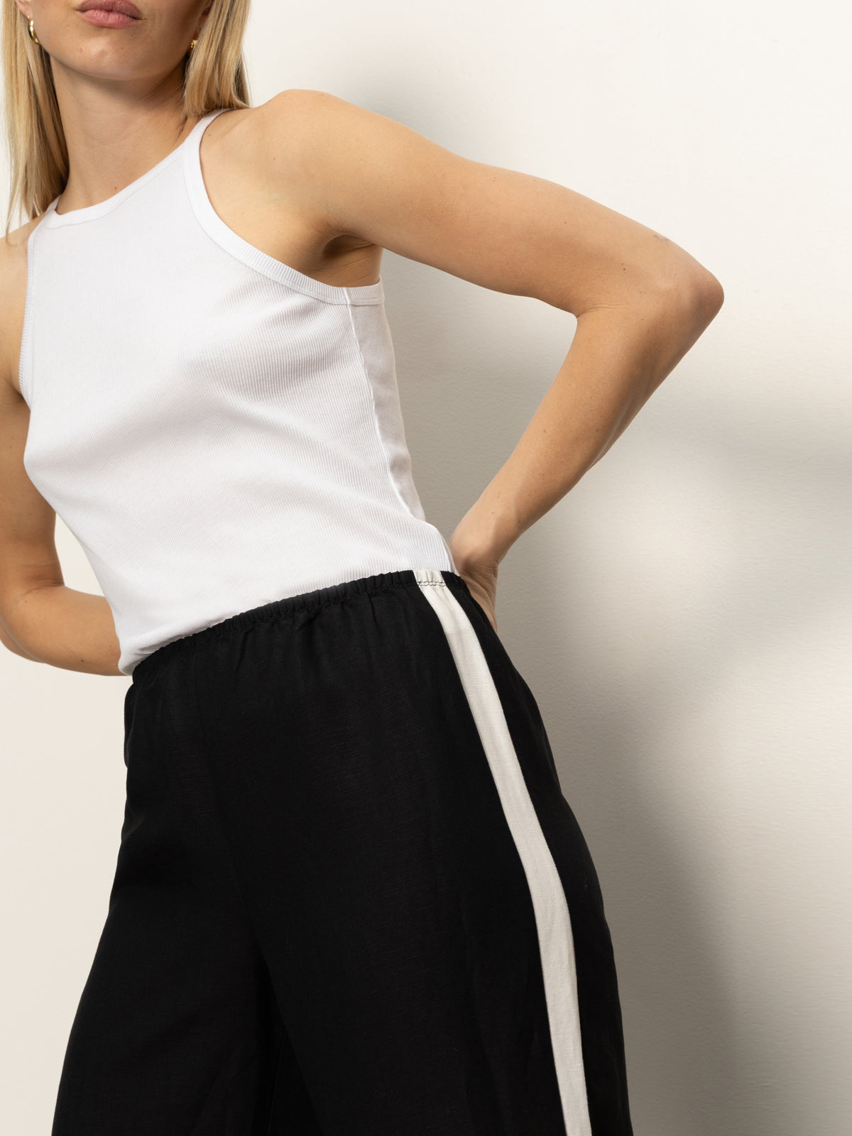 A woman models Sanctuary Clothing’s casual track pant in black with a white chalk stripe, pairing it with a sleeveless white tank top and posing against a plain light background, one hand on her hip and her head partially out of frame.