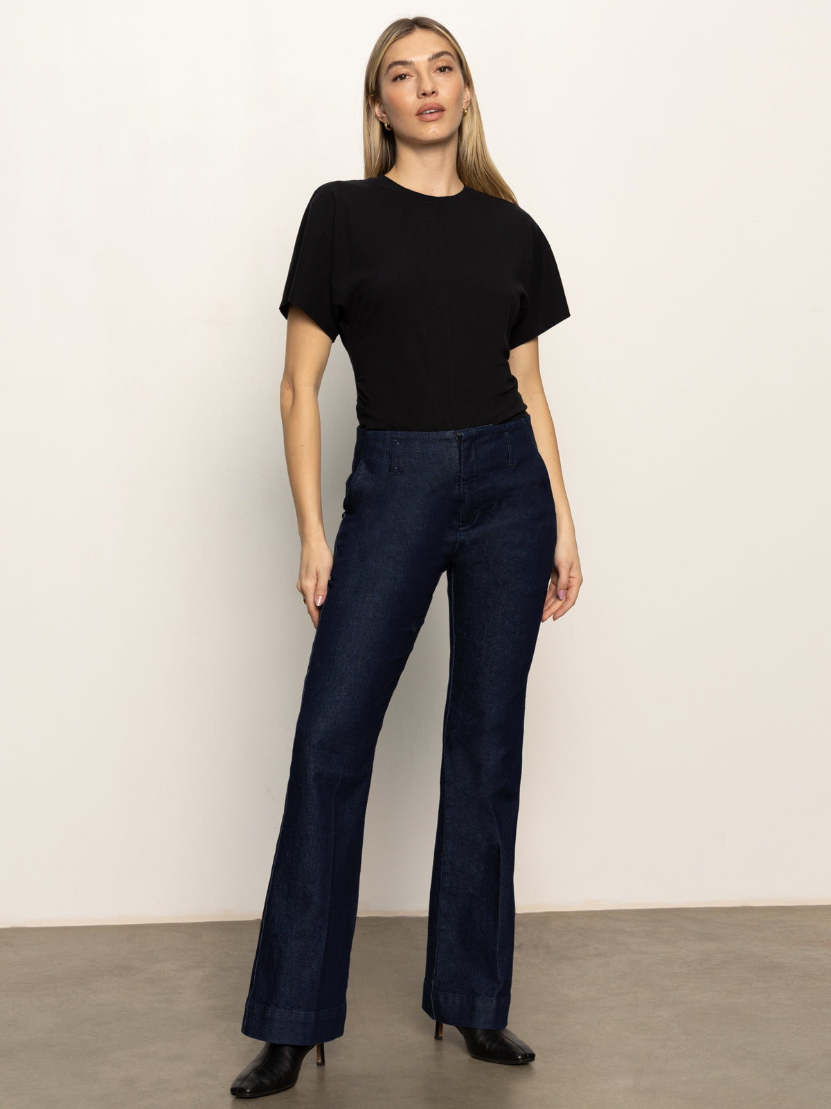 A woman poses confidently against a light wall, wearing the Sanctuary Clothing denim stunner jean plot twist—dark blue high-waisted wide-leg jeans—with a fitted black short-sleeve top and black heeled boots. Her arms are relaxed by her sides.