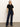 A woman poses confidently against a light wall, wearing the Sanctuary Clothing denim stunner jean plot twist—dark blue high-waisted wide-leg jeans—with a fitted black short-sleeve top and black heeled boots. Her arms are relaxed by her sides.