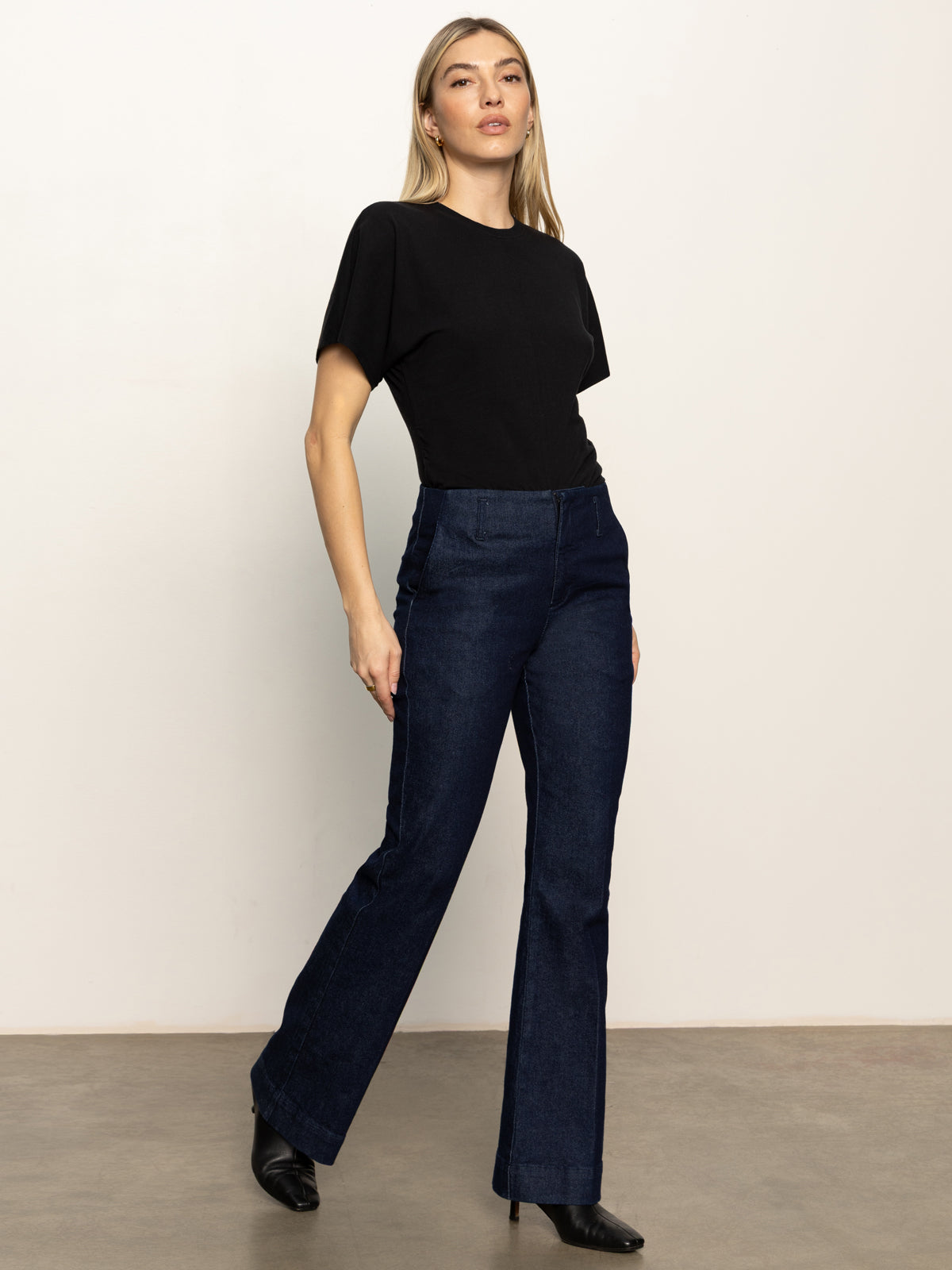 A woman with long, straight blonde hair and a confident look models Sanctuary Clothing’s “denim stunner jean plot twist,” a dark blue high-waisted wide-leg style, paired with a black short-sleeve top and black heeled boots against a plain background.