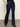 A person models the Sanctuary Clothing denim stunner jean plot twist, showcasing its high-waisted, dark blue design paired with black heeled boots and one hand in their pocket; only the lower body is visible against a plain background.