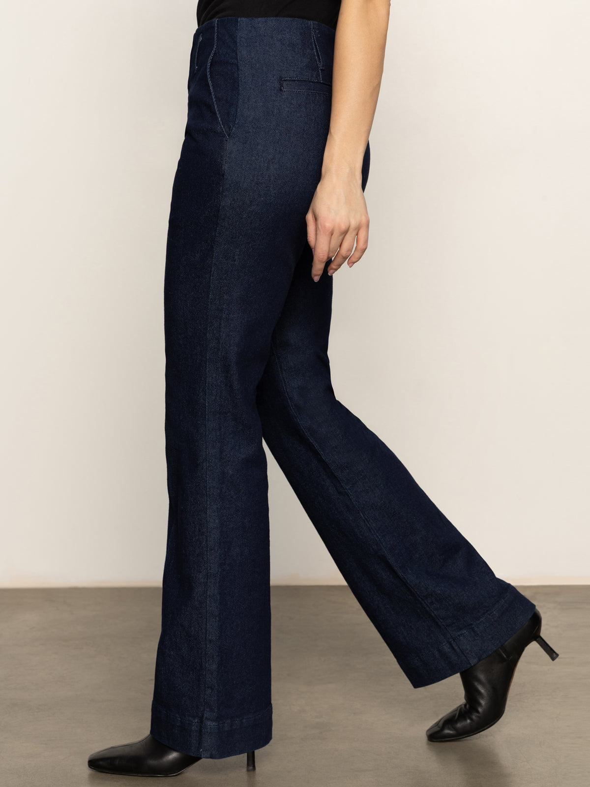 A person wearing Sanctuary Clothing's denim stunner jean plot twist and black heeled shoes walks indoors on a gray floor, with only the lower half of their body visible against a plain light-colored wall.