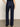 A person is shown from the back wearing Sanctuary Clothing’s dark blue denim stunner jean plot twist and black heels, standing on a gray floor with their left arm slightly bent and hand resting by their side.