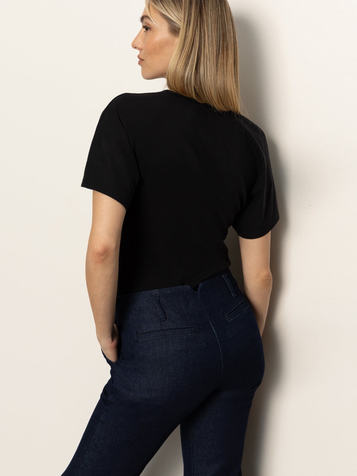 A woman with straight blonde hair stands facing away from the camera, wearing a black short-sleeve top and Sanctuary Clothing’s denim stunner jean plot twist against a light-colored wall.