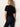 A woman with straight blonde hair stands facing away from the camera, wearing a black short-sleeve top and Sanctuary Clothing’s denim stunner jean plot twist against a light-colored wall.