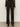 A person wears Sanctuary Clothing's the stunner pant luster stripe€�a black, high-waisted, straight-leg pant with subtle glittery pinstripes€�paired with a sheer black top and black shoes, standing against a plain light background.