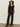 A woman with curly hair stands against a plain wall, wearing Sanctuary Clothing„¢s the stunner pant luster stripe, paired with a sheer black ruffled blouse and black heeled boots, one hand in her pocket.