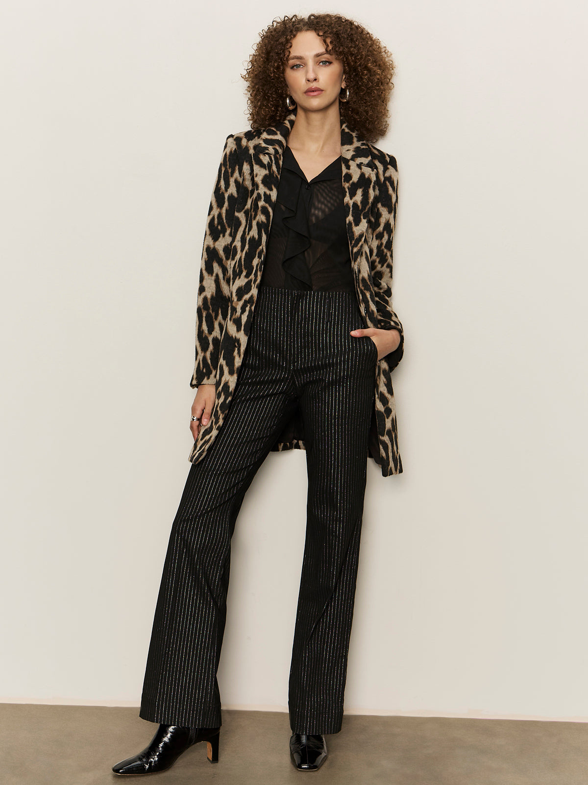 A woman with curly hair wears the Stunner Pant Luster Stripe by Sanctuary Clothing, paired with a leopard print blazer, black blouse, and black heeled boots. She stands against a plain light-colored wall with one hand in her pocket.